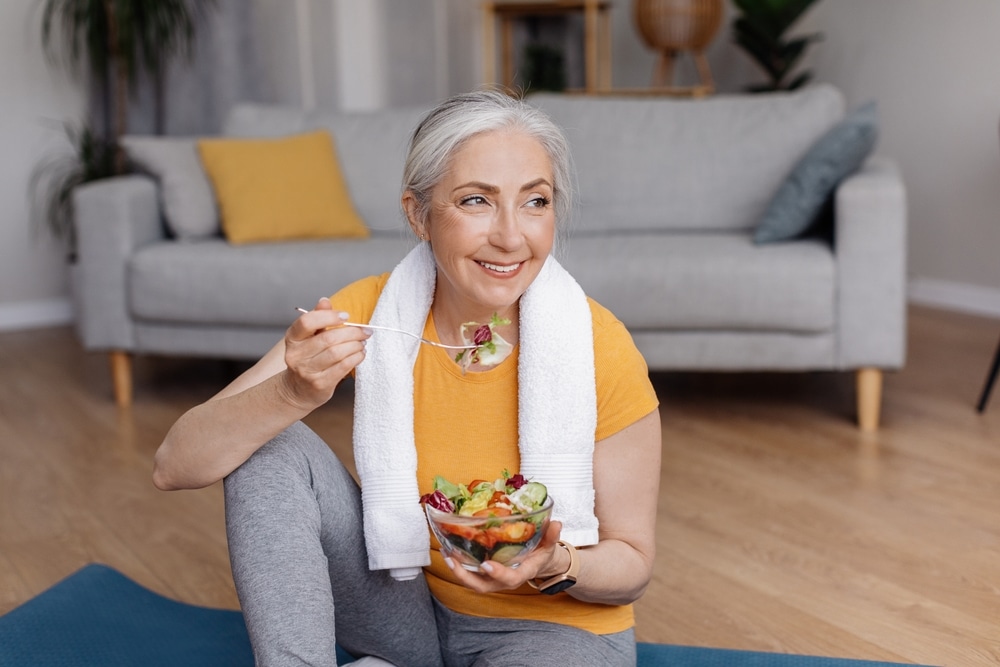Smiling older woman sitting on a yoga mat, eating a fresh salad, representing healthy nutrition and joint support for active aging.