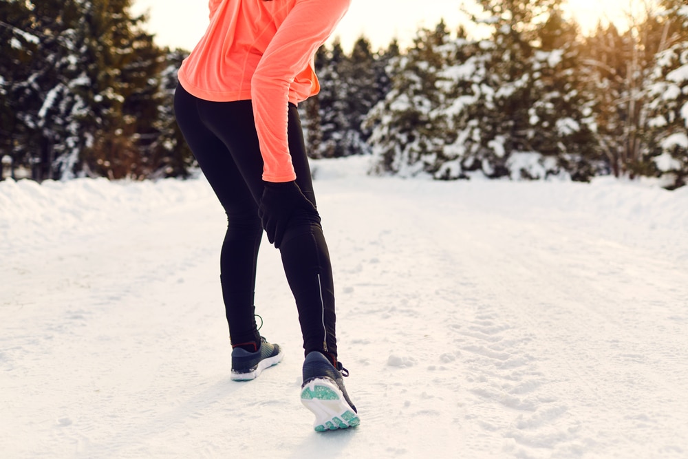 Woman out in wintery woods, holding the back of her knee after getting injured on her winter run