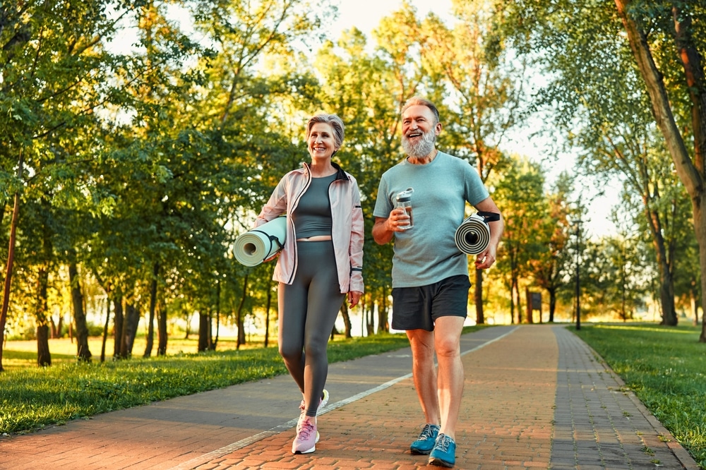 older healthy couple walking outside in a park and holding yoga mats.