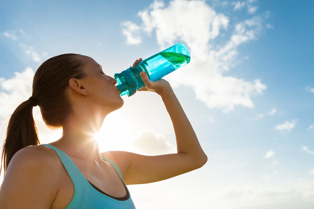 Woman drinking water outdoors in the sun, representing hydration during summer activity to help reduce muscle cramping.